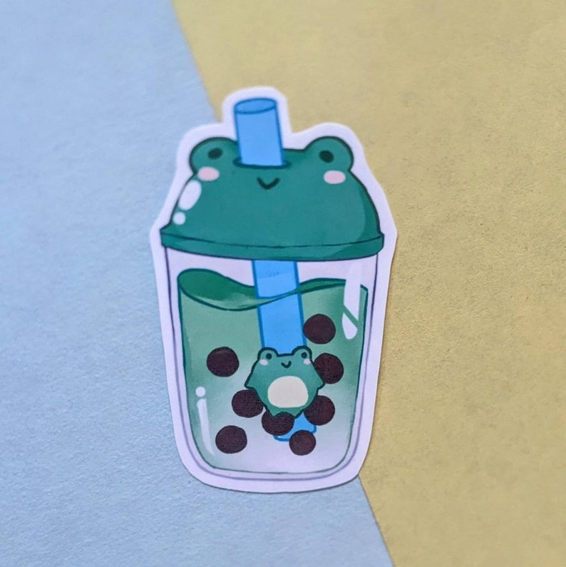 May include: A green frog-shaped lid on a clear plastic cup with a blue straw. The cup is filled with green liquid and black boba pearls. A small frog is inside the cup.