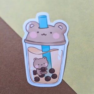 May include: A brown bear-shaped sticker with a blue straw, sitting inside a clear cup with brown boba pearls and a light brown liquid.