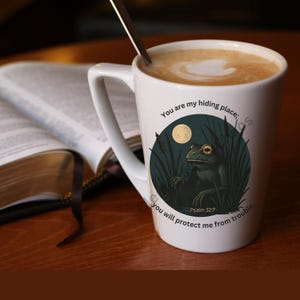 May include: White ceramic mug with a frog illustration, a full moon, and text that reads "You are my hiding place; you will protect me from trouble." A spoon rests inside the mug, which is filled with a light brown beverage.