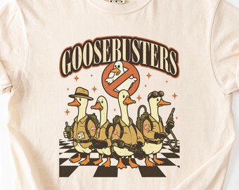 May include: Cream-colored t-shirt featuring the text "GOOSEBUSTERS" above a graphic of four cartoon geese dressed as Ghostbusters. The geese are standing on a black and white checkered pattern.