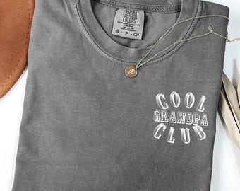 May include: A gray Comfort Colors t-shirt with the text "COOL GRANDPA CLUB" in white. A silver necklace with a circular pendant rests on the shirt. A brown hat and dried leaves are in the background.