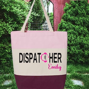 May include: Pink and cream tote bag with a burgundy bottom. The bag has a black and white graphic that says "Dispatcher" with a pink headset icon and the name "Emily" underneath.