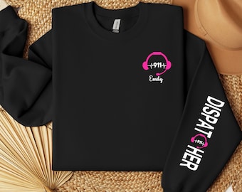 Op de afbeelding: Zwart sweatshirt met een roze afbeelding van een headset met een hartslag en de tekst "911" en "Emily". De rechtermouw heeft de tekst "DISPATCHER" met een roze afbeelding van een headset met een hartslag en de tekst "911".