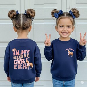 May include: Navy blue sweatshirt with the text "IN MY HORSE GIRL ERA" and a horse graphic on the back. The front of the sweatshirt has the name "Samantha" and a horse graphic. The sweatshirt is worn by a young girl.