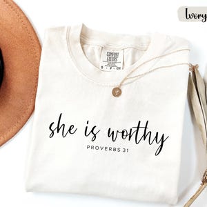 May include: Ivory-colored t-shirt with the words "she is worthy" in black script, and "PROVERBS 31" below. A gold necklace with a pendant rests on the shirt. A brown hat and dried plant are in the background.
