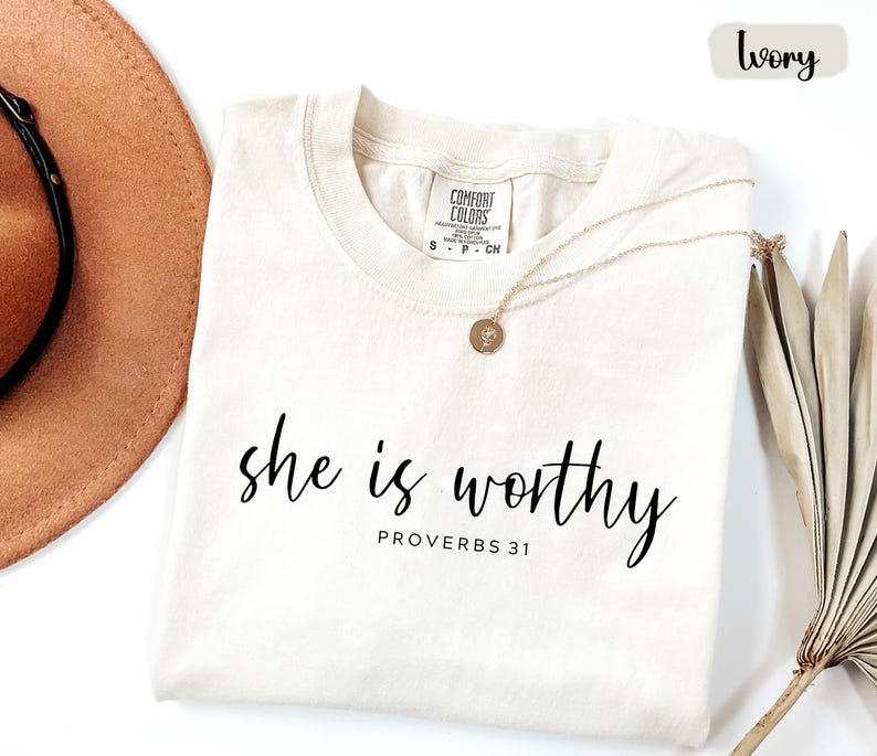 May include: Ivory-colored t-shirt with the words "she is worthy" in black script, and "PROVERBS 31" below. A gold necklace with a pendant rests on the shirt. A brown hat and dried plant are in the background.