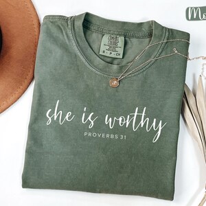 May include: A moss green t-shirt with the words "she is worthy" and "PROVERBS 31" printed in white script. A gold necklace with a pendant rests on the shirt. A brown hat and dried palm leaves are in the background.
