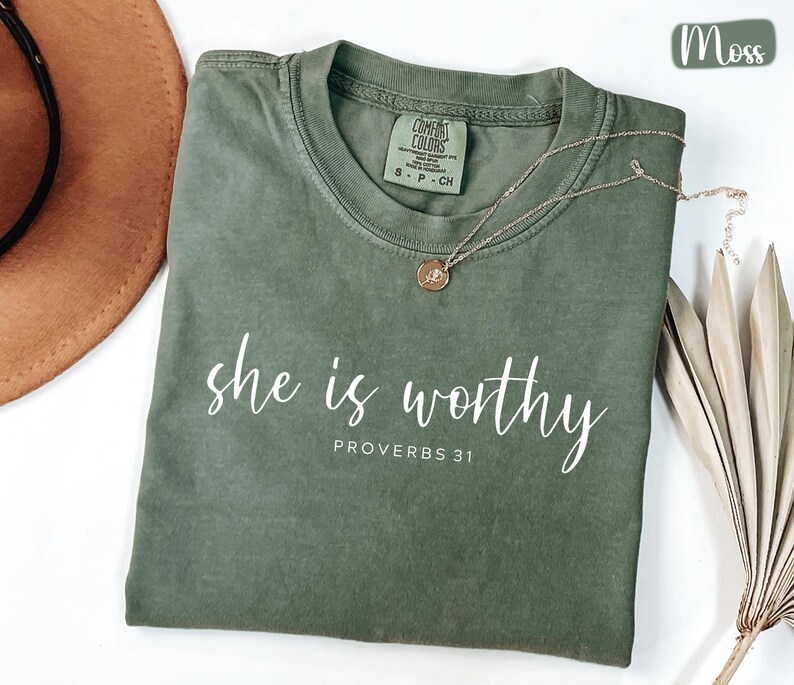 May include: A moss green t-shirt with the words "she is worthy" and "PROVERBS 31" printed in white script. A gold necklace with a pendant rests on the shirt. A brown hat and dried palm leaves are in the background.