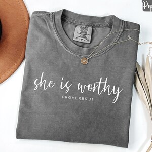 May include: A gray t-shirt with the words "she is worthy" and "Proverbs 31" printed in white. A gold necklace with a pendant is draped on the shirt. The shirt is folded on a white surface.