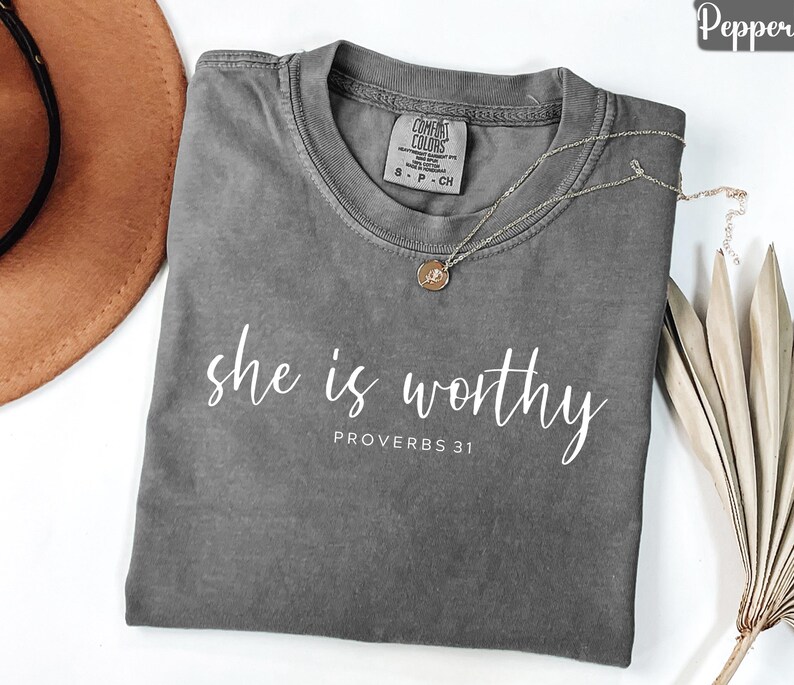 May include: A gray t-shirt with the words "she is worthy" and "Proverbs 31" printed in white. A gold necklace with a pendant is draped on the shirt. The shirt is folded on a white surface.