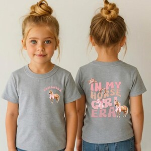May include: Two children wearing gray t-shirts with horse-themed designs. The front of one shirt says "Samantha" above a horse with a pink bow. The back of the other shirt says "In My Horse Girl Era" with horse graphics.