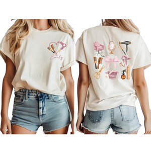 Op de afbeelding: Cr&egrave;mekleurig T-shirt met een hartvormig ontwerp met een f&ouml;hn, kam en schaar, met de naam "Emily". De achterkant van het shirt toont diverse salon gereedschappen en accessoires, waaronder een stoel, spiegel en f&ouml;hn.