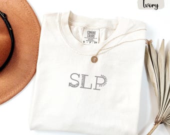May include: Ivory-colored t-shirt with the letters "SLP" in gray, with "SPEECH" and "PATHOLOGIST" circling the letters. A gold necklace with a pendant rests on the shirt. The shirt is labeled "COMFORT COLORS".