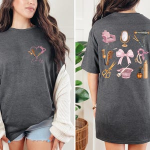 Op de afbeelding: Donkergrijs t-shirt met een hartontwerp met salon gereedschap en de naam "Emily" op de voorkant. De achterkant van het shirt toont diverse salon gereedschappen, waaronder een stoel, spiegel, schaar en f&ouml;hn. Een roze strik is ook afgebeeld.
