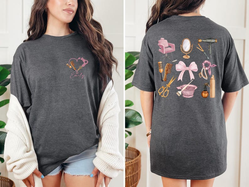 Op de afbeelding: Donkergrijs t-shirt met een hartontwerp met salon gereedschap en de naam "Emily" op de voorkant. De achterkant van het shirt toont diverse salon gereedschappen, waaronder een stoel, spiegel, schaar en f&ouml;hn. Een roze strik is ook afgebeeld.