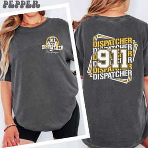May include: Dark grey t-shirt with "911 Dispatcher" text and a graphic on the front and back. The front graphic includes a badge and the name "Mackenzie". The back features a larger design with "911" and "Dispatcher" in yellow and white.