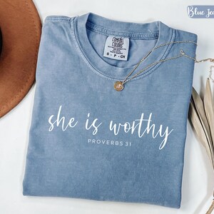 May include: A blue jean colored t-shirt with the words "she is worthy" and "Proverbs 31" printed in white. A gold necklace with a pendant rests on the shirt. The shirt is folded and displayed with a brown hat and dried plant.