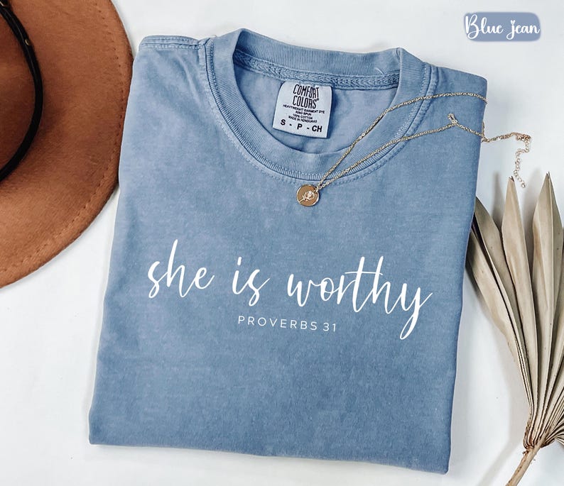 May include: A blue jean colored t-shirt with the words "she is worthy" and "Proverbs 31" printed in white. A gold necklace with a pendant rests on the shirt. The shirt is folded and displayed with a brown hat and dried plant.