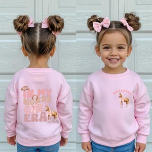 May include: A pink sweatshirt with the text "IN MY HORSE GIRL ERA" and a horse graphic on the back. The front of the sweatshirt has the name "Samantha" and a horse graphic. The child is wearing blue jeans.