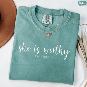 May include: A light green t-shirt with the words "she is worthy" and "PROVERBS 31" printed in white. A gold necklace with a small pendant rests on the shirt. The shirt is folded and has a tag.