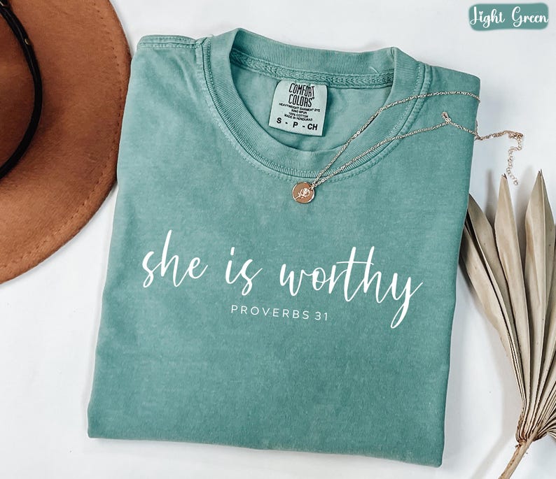May include: A light green t-shirt with the words "she is worthy" and "PROVERBS 31" printed in white. A gold necklace with a small pendant rests on the shirt. The shirt is folded and has a tag.
