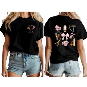 Op de afbeelding: Zwart t-shirt met een roze hartontwerp en haarstylingtools, plus de naam "Emily" op de voorkant. De achterkant toont salon tools zoals een stoel, spiegel en f&ouml;hn, in roze en goud.