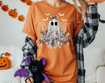 May include: Orange t-shirt with a ghost design, bow, lace, and floral details. The ghost is surrounded by spiderwebs and bats, ideal for Halloween. The shirt is a comfortable, casual style.