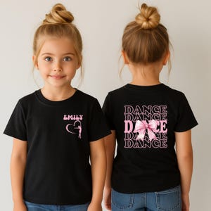 May include: Black t-shirt with pink text and graphics. The front features the name "EMILY" above a heart-shaped design with a ballerina. The back of the shirt displays the word "DANCE" repeated four times with a pink bow graphic.