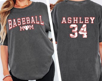 May include: Dark gray t-shirt with the words "BASEBALL MOM" in red and white lettering on the front, with a heart in place of the "O". The back of the shirt has "ASHLEY" and the number "34" in red and white lettering.