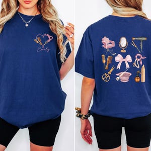 Op de afbeelding: Marineblauw T-shirt met een hartvormig ontwerp met haarstylingtools en de naam "Emily". De achterkant van het shirt toont diverse salon tools. Een stijlvol t-shirt voor kappers.