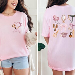 Op de afbeelding: Lichtroze t-shirt met een hartvormig ontwerp met haarstylingtools en de naam "Emily". De achterkant van het shirt toont diverse salon tools en accessoires, waaronder een stoel, spiegel en schaar.