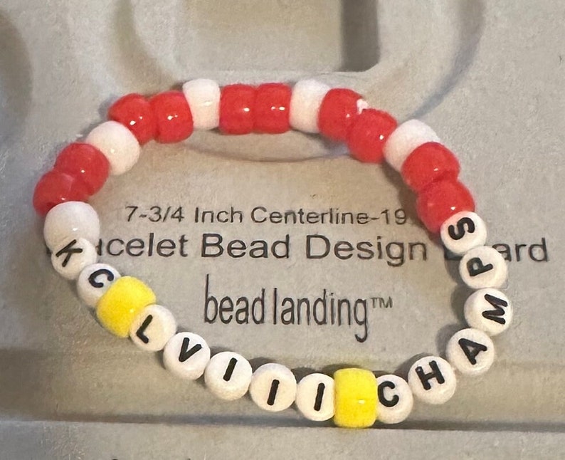 Puede incluir: Un brazalete de cuentas blancas y rojas con la palabra "CLEVELAND CHAMPS" escrita en letras negras sobre cuentas blancas. El brazalete tiene una circunferencia de 19,7 cm y est&aacute; hecho por bead landing.