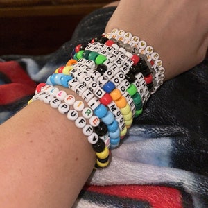 Puede incluir: Una mu&ntilde;eca est&aacute; adornada con una pila de pulseras de cuentas de colores, cada una con una palabra diferente escrita en letras blancas. Las palabras incluyen "Dream", "Meme", "Ranger", "Upside", "Duck", "Bloody", "Yemo", "Tid", "Fire", "Puff", "Moon", "Tho", "Do", "Fe" y "Tell".