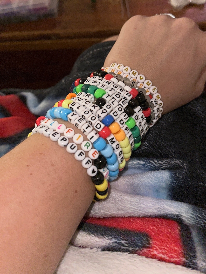 Puede incluir: Una mu&ntilde;eca est&aacute; adornada con una pila de pulseras de cuentas de colores, cada una con una palabra diferente escrita en letras blancas. Las palabras incluyen "Dream", "Meme", "Ranger", "Upside", "Duck", "Bloody", "Yemo", "Tid", "Fire", "Puff", "Moon", "Tho", "Do", "Fe" y "Tell".