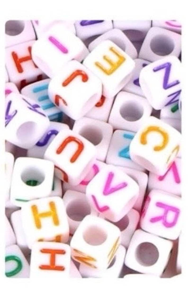 Puede incluir: Un mont&oacute;n de cuentas de letras de pl&aacute;stico blanco con letras de colores. Las cuentas miden aproximadamente 1 cm y tienen un agujero en el centro para enhebrar.