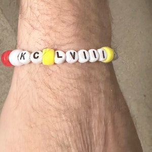 Puede incluir: Un brazalete de cuentas blancas con las letras "KCLVIII" en negro. Las cuentas alternan entre amarillo y rojo.