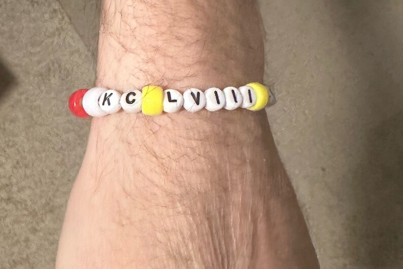 Puede incluir: Un brazalete de cuentas blancas con las letras "KCLVIII" en negro. Las cuentas alternan entre amarillo y rojo.
