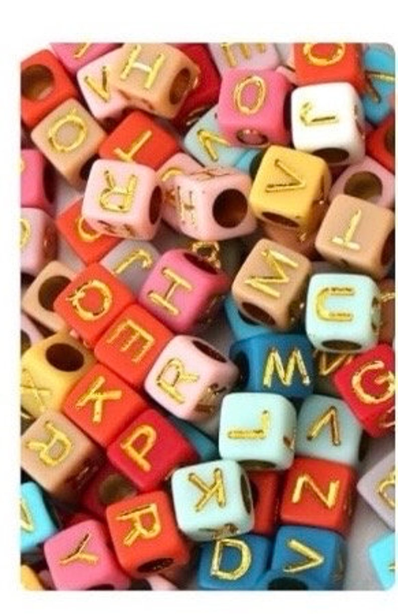 Puede incluir: Primer plano de cuentas de letras del alfabeto de colores. Las cuentas son de pl&aacute;stico y tienen un acabado mate. Las letras son doradas.