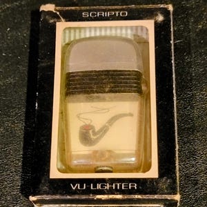 May include: A vintage Scripto VU-Lighter in its original box. The lighter has a clear plastic body with a black and gray top. A black pipe with a red bowl is printed on the front. The box is black with the Scripto and VU-Lighter text.