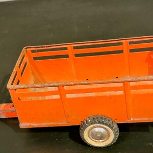 May include: An orange toy trailer with black horizontal stripes on the sides and back. The trailer has a single tire with a white rim and a metal hitch. The toy is likely vintage and shows signs of wear.