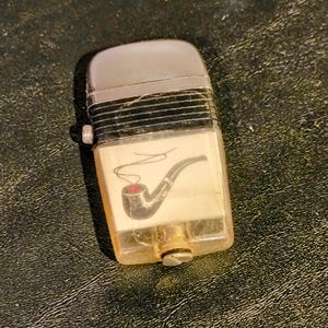 May include: A vintage lighter with a silver top and a clear plastic body. The lighter features a graphic of a smoking pipe with a red bowl. The lighter is rectangular and has a small screw at the bottom.