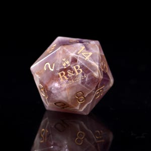 May include: A purple and white amethyst gemstone dice with gold numbers and the inscription "R&B 15.24.2024".