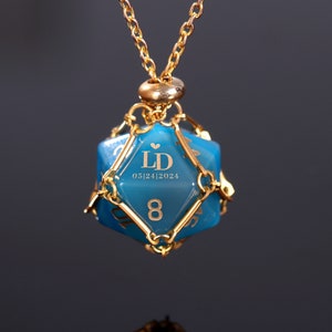 May include: A gold necklace with a blue, faceted, dice-shaped pendant. The pendant has gold wire accents and is engraved with the initials "LD" and the date "05/24/2024".