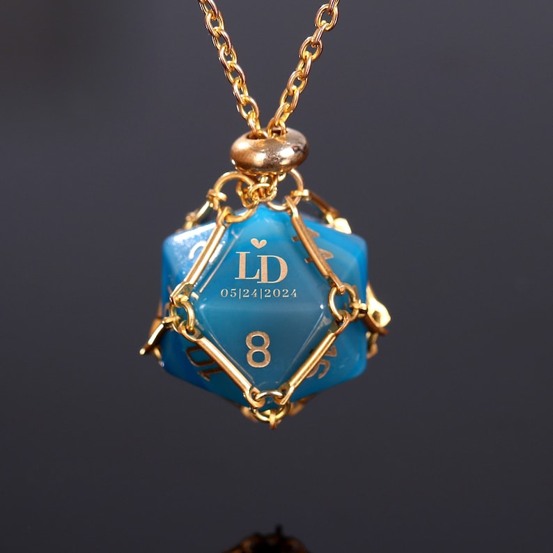 May include: A gold necklace with a blue, faceted, dice-shaped pendant. The pendant has gold wire accents and is engraved with the initials "LD" and the date "05/24/2024".