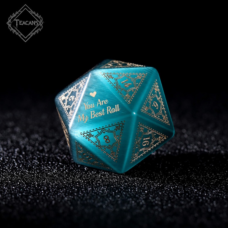 May include: A teal, multi-sided die with gold-colored numbers and decorative patterns. The text "You Are My Best Roll" is featured on one side, along with a heart symbol. The die is designed for tabletop games.