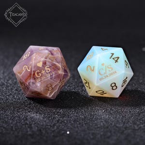 May include: Two personalized d20 dice. One is amethyst-colored with gold numbers and initials "G+S" and the date "05.24.2024". The other is opalescent with gold numbers and initials "C/S" and the date "05.24.2024".