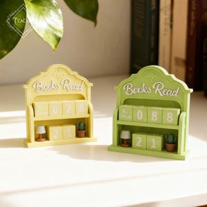 Op de afbeelding: Twee decoratieve houten kalenders met boeken als thema. De ene is geel, de andere groen. Elk heeft de tekst "Books Read" bovenaan. De gele kalender toont "028" en "21". De groene kalender toont "088" en "21".