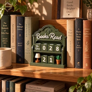 May include: A dark green wooden desk calendar with the text Books Read, featuring four rotatable blocks displaying the numbers 0, 2, 8, 2, and 1, along with a small decorative lamp and a potted cactus figurine, positioned between several hardcover books.