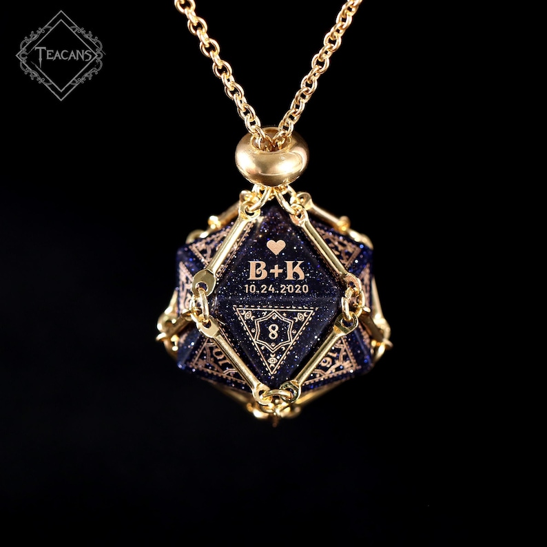 May include: A gold-colored necklace with a pendant. The pendant is a dark blue, multi-faceted shape with gold-colored accents. The pendant has the initials "B+K", a heart, the date "10.24.2020", and the number "8" engraved on it. The necklace is on a black background.