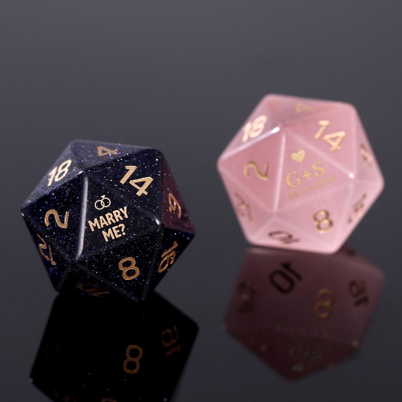 May include: Two 20-sided dice, one blue with gold glitter and the other pink. The blue die has the words "Marry Me?" and a ring symbol. The pink die has the initials "G+S" and a heart symbol.